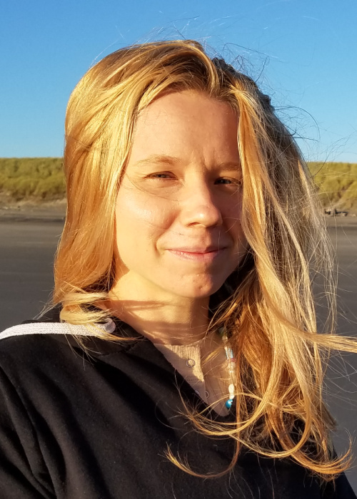 a photograph of Julia. She is a 30 year old white woman who is smiling and sunlit on the beach. Her hair is blonde and blowing in the wind about her.